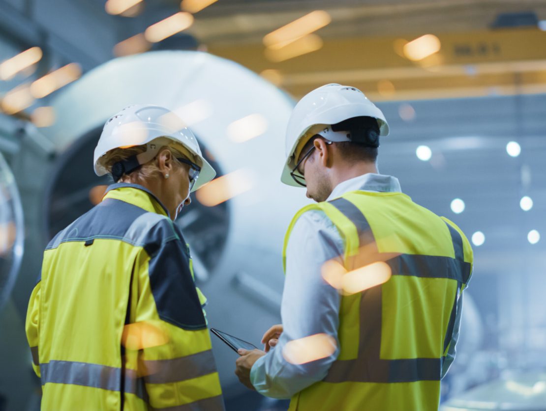 Two Heavy Industry Engineers Stand in Pipe Manufacturing Factory, Use Digital Tablet Computer, Have Discussion. Construction of Oil, Gas and Fuels Transport Pipeline. Back View Sparks Flying